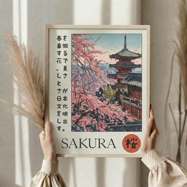 May include: Person holding a framed poster featuring a traditional Japanese pagoda temple surrounded by pink cherry blossom trees in an illustrated vintage style. The artwork includes Japanese text and "SAKURA" with a red circle symbol below. Light wooden frame, held against neutral curtained background with dried wheat stems visible to the left.