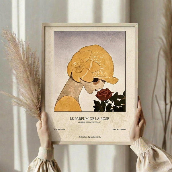 May include: Hands holding a framed vintage-style art print featuring a woman in profile wearing a golden turban-style headwrap, smelling a dark red rose. The illustration uses warm earth tones with French text below reading "LE PARFUM DE LA ROSE." Dried wheat stalks are visible to the left, and the scene is set against soft natural lighting near a window with flowing curtains.