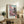 May include: Framed poster featuring pink cherry blossoms and traditional Japanese pagoda with vertical Japanese text and "SAKURA" at bottom, displayed on rustic wooden console table alongside potted fern, ceramic vases, and books in neutral-toned living room with beige chair