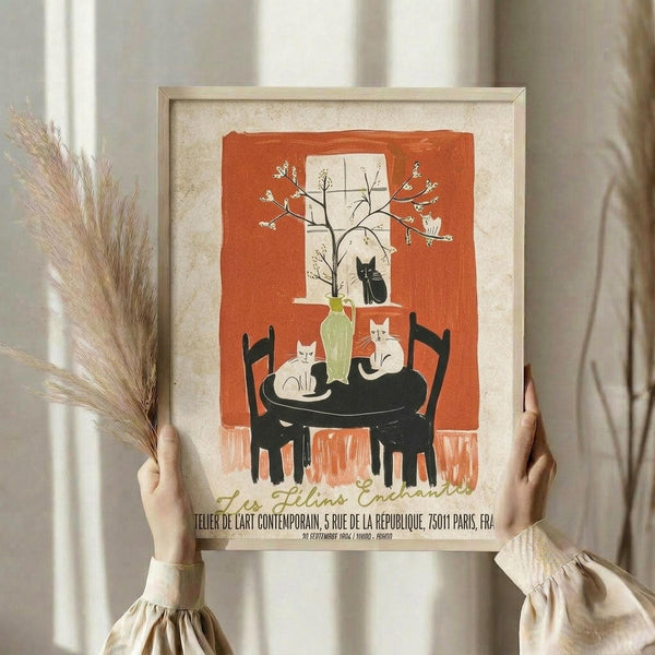 May include: Person holding framed vintage-style poster featuring orange and cream illustration of cats around black table with green vase and flowering branches, displayed in neutral room setting with dried pampas grass and soft natural lighting