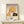 May include: Framed vintage-style poster showing woman in golden yellow cloche hat smelling deep red rose, with French text "Le Parfum de la Rose" below illustration. Art deco inspired design in warm earth tones displayed on light wooden shelf next to small wrapped gift with evergreen sprig. Natural wood frame against cream wall.