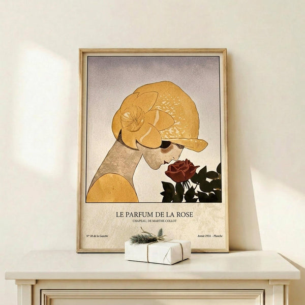 May include: Framed vintage-style poster showing woman in golden yellow cloche hat smelling deep red rose, with French text "Le Parfum de la Rose" below illustration. Art deco inspired design in warm earth tones displayed on light wooden shelf next to small wrapped gift with evergreen sprig. Natural wood frame against cream wall.