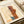 May include: Paul Klee abstract artwork print from 1924 displayed on cream-colored paper in an open book or portfolio, alongside rolled white papers and beige ribbon. The modernist composition features geometric shapes in warm browns, oranges, and blue-green tones with linear sketched elements against a neutral background.