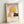 May include: Framed vintage-style art print leaning against white wall showing woman in golden yellow cloche hat smelling red rose, with French text "Le Parfum de la Rose" below illustration. Light wood frame, cream background with subtle texture, warm lighting casting soft shadows.