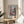 May include: Framed poster displaying Japanese pagoda temple with pink cherry blossoms in vintage travel poster style, featuring Japanese text and "SAKURA" with red circular logo. Print sits on light wood round side table next to beige upholstered chair with white pillow. Soft natural lighting creates shadows on cream wall, with green plant leaves visible in corner.