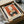 May include: Vintage-style poster print featuring three cats sitting around a small black table with a potted plant, set against a rust orange and cream background with delicate botanical illustrations. The framed artwork is shown in a cardboard shipping box with protective wrapping paper, photographed on a white surface next to what appears to be a dark glass object.