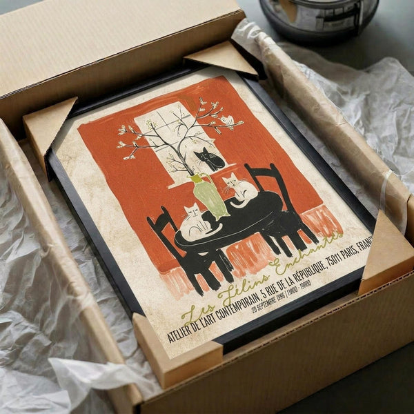 May include: Vintage-style poster print featuring three cats sitting around a small black table with a potted plant, set against a rust orange and cream background with delicate botanical illustrations. The framed artwork is shown in a cardboard shipping box with protective wrapping paper, photographed on a white surface next to what appears to be a dark glass object.