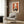 May include: Vintage-style French cat poster with orange background featuring black and white cats around a dark table with flowering branches in a vase, displayed in white frame on wood-paneled wall above modern beige armchair with geometric pillow, brass floor lamp and side table with open book nearby in contemporary living room setting.