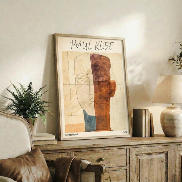 May include: Framed Paul Klee abstract art print displayed on rustic wooden sideboard in warm, neutral-toned living room. The artwork features geometric shapes in orange, brown, and blue tones with linear elements. Scene includes fern plant, ceramic vase, stacked books, and beige upholstered chair, creating a cozy modern home interior setting.