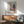 May include: Framed poster showing Japanese pagoda temple with pink cherry blossoms, Japanese text, and "SAKURA" title with red circular stamp. Displayed above mid-century modern wood credenza with white ceramic lamp. Sage green upholstered armchair with wooden arms positioned nearby. Natural sunlight creates diagonal shadows on white wall. Vintage travel poster aesthetic with muted pink, blue, and brown color palette.