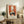 May include: Framed vintage-style poster with orange background featuring black and white cats around a table, with a flowering branch above. The artwork is displayed on a rustic wooden console alongside books, a small plant, and ceramic vases in a cozy living room setting with neutral tones.