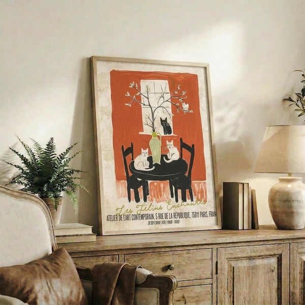 May include: Framed vintage-style poster with orange background featuring black and white cats around a table, with a flowering branch above. The artwork is displayed on a rustic wooden console alongside books, a small plant, and ceramic vases in a cozy living room setting with neutral tones.