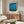 May include: Blue abstract art poster with red and orange accents hanging above wooden sideboard in minimalist living room. White curved chair and glass coffee table with colorful artwork visible. Warm wood tones throughout modern interior with natural lighting and neutral walls.