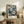 May include: Framed floral art print with white and blue cosmos flowers on dark navy background, displayed on rustic wooden sideboard next to fern plant, ceramic vase, and books in neutral-toned living room setting with beige upholstered furniture.