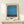 May include: Framed abstract art print with horizontal bands of blue watercolor paint and orange-red accents at edges, displayed on light wood shelf against white wall. Small wrapped gift box sits in front of the natural wood frame. The artwork features soft, blended brushstrokes in various blue tones creating layered rectangular forms typical of color field painting style.