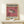 May include: Framed exhibition poster with pink background featuring detailed pufferfish illustration, displayed on light wood shelf against cream wall. Poster shows Japanese and English text including "1974" and "The Ureki Gallery" with San Francisco Japantown address. Small wrapped gift box with twine sits in front of thin gold frame.