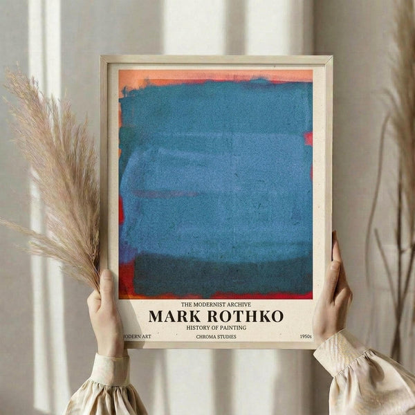 May include: Person in cream blouse holding framed poster featuring abstract blue watercolor painting with coral-orange accents, displayed in light wooden frame. Dried wheat stalks visible to left, soft natural lighting against neutral curtain backdrop. Poster shows museum-style text below artwork referencing modernist archive and painting studies from 1950s.
