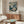 May include: Framed floral art print displayed on wooden sideboard in modern living room. Print features cream and blue flowers on dark navy background with "YAYOI KUSAMA" and Japanese text. Room has white curved chair, brass floor lamp, glass-top coffee table, and neutral walls with crown molding.
