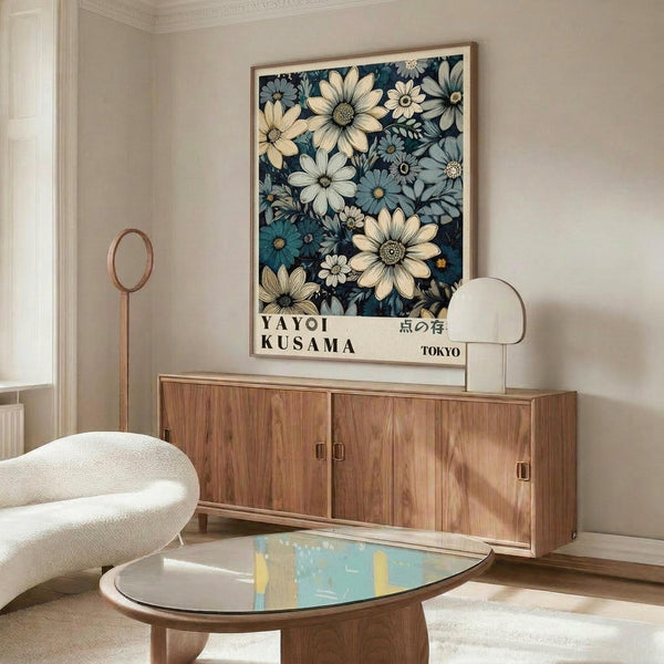May include: Framed floral art print displayed on wooden sideboard in modern living room. Print features cream and blue flowers on dark navy background with "YAYOI KUSAMA" and Japanese text. Room has white curved chair, brass floor lamp, glass-top coffee table, and neutral walls with crown molding.