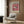 May include: Pink and cream exhibition poster with pufferfish illustration hanging on wood-paneled wall above beige upholstered armchair with geometric pillow. Modern floor lamp with brass finish and round marble base stands beside chair. Dark round side table with open book visible on left. Warm wood textures throughout minimalist interior setting.