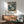 May include: Framed floral art print hanging above mid-century modern wooden credenza and sage green upholstered chair. Print features cream and blue flowers on dark background with "YAYOI KUSAMA" and Japanese text at bottom. Warm sunlight creates geometric shadows on white wall. Small white ceramic lamp sits on credenza next to round wooden side table.