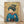 May include: Framed poster print leaning against white wall on light wood floor. Artwork shows Japanese woman in traditional blue kimono with geometric patterns and red collar, elaborate dark hair with ornate pins, holding thin sticks or chopsticks. Background features golden yellow tones with delicate cherry blossom branches. Art style resembles vintage Japanese woodblock prints with detailed line work and muted color palette. Natural wood frame visible around edges.
