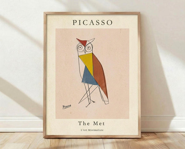 May include: Framed poster print leaning against white wall on light wood floor. Features geometric owl illustration in Picasso-inspired style with colorful triangular shapes in yellow, blue, and rust red. Simple line drawing with large circular eyes and thin legs. Text reads "PICASSO" at top and "The Met L'Art Minimaliste" at bottom. Light wood frame with cream matting on textured beige background.