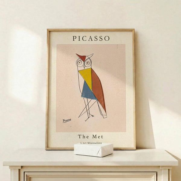 May include: Framed poster print featuring geometric owl artwork in Picasso's minimalist style, displayed on light wood shelf against cream wall. Owl composed of colorful angular shapes - red triangular head accent, yellow and blue geometric body sections, orange-brown wing, thin black line legs. Natural wood frame with cream matting. Small white geometric object sits beside frame on shelf. Soft natural lighting creates gentle shadows.