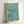 May include: Framed poster print leaning against white wall showing three swimmers diving through turquoise blue water with white splash effects. Features motivational text "Finish the race no matter what it takes" in English and vertical Japanese characters along right side. Light wood frame, watercolor-style artwork with minimalist aesthetic.