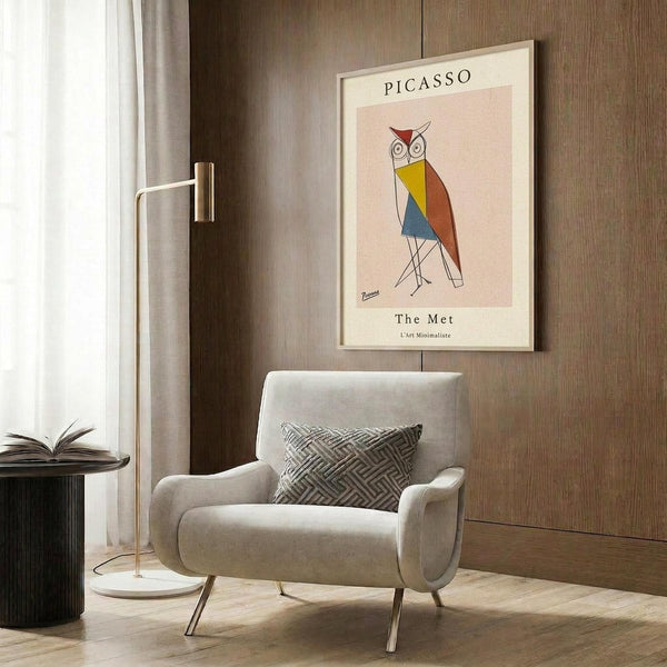 May include: Framed poster featuring a geometric owl in Picasso's minimalist style with colorful angular shapes in yellow, blue, orange and red against a pink background. The print hangs on wood-paneled wall above a cream upholstered armchair with patterned pillow. Modern reading nook includes brass floor lamp and side table with open book.