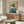 May include: Japanese woodblock print-style artwork showing a geisha in blue kimono with red collar against golden cherry blossom background, displayed above a mid-century walnut credenza with brass handles. Modern living room setting features white curved chair, oval glass coffee table, brass floor lamp with circular mirror detail, and neutral walls with natural lighting.