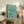 May include: Framed poster with turquoise blue background showing three orange-toned swimmers in diving poses, displayed on rustic wooden sideboard alongside potted plants, books, and ceramic vases in neutral-toned living room setting with cream chair visible.