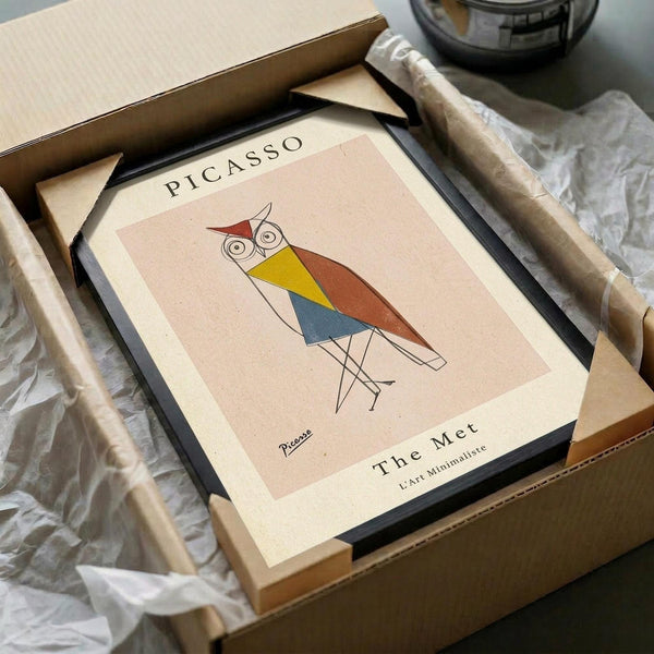 May include: Framed minimalist owl print in geometric style with red, yellow, and blue triangular shapes on cream background, packaged in brown cardboard box with protective tissue paper, displayed on white fabric surface alongside wooden tubes and glass object