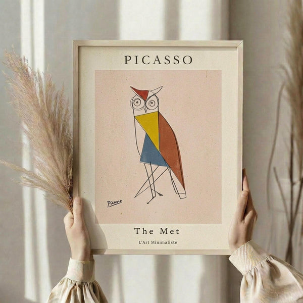 May include: Hands holding a framed poster featuring a geometric owl illustration in Picasso's minimalist style, with colorful triangular shapes in yellow, blue, and rust red against a cream background. The print shows "PICASSO" at top and "The Met L'Art Minimaliste" at bottom. Person wears beige clothing and holds dried pampas grass, photographed against white curtained window.