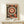 May include: Framed abstract art print displayed on light wood console table against white wall. Design features large central mandala with concentric circles in orange, cream and black, surrounded by smaller circular and organic patterns. Dark background with geometric and dotted elements throughout. Small wrapped gift box with greenery sits nearby. Light wood frame complements minimalist modern setting.