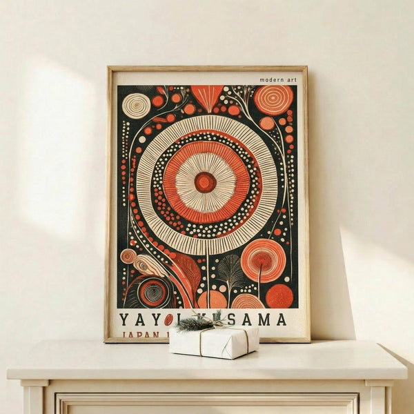 May include: Framed abstract art print displayed on light wood console table against white wall. Design features large central mandala with concentric circles in orange, cream and black, surrounded by smaller circular and organic patterns. Dark background with geometric and dotted elements throughout. Small wrapped gift box with greenery sits nearby. Light wood frame complements minimalist modern setting.