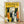 May include: Framed vintage-style cat illustration poster leaning against white wall on wooden floor. Yellow background features collection of stylized cats in various colors - black, orange, white, gray, striped - with geometric folk art design. Natural wood frame surrounds the print in bright room setting with natural lighting casting shadows on textured white wall.