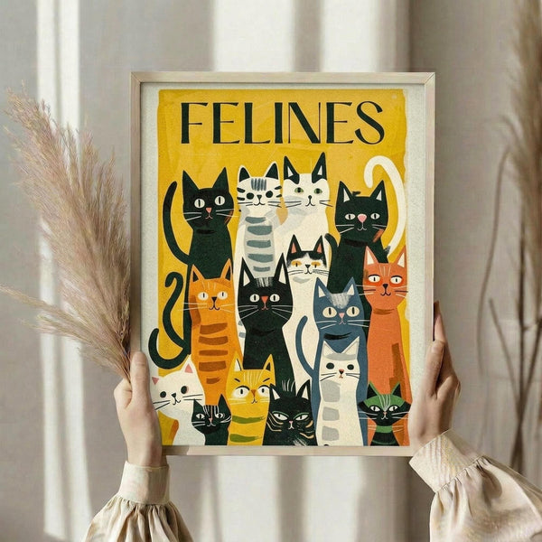May include: Person holding framed cat poster with dried pampas grass. Yellow background features illustrated cats in various colors - black, orange, striped, and white - arranged in group formation. Bold "FELINES" text at top. Light wood frame against white curtained window background.