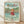 May include: Framed Studio Ghibli Ponyo poster showing red-haired child character splashing joyfully in turquoise ocean waves with arms raised, coastal town and green hills in background, Japanese and English text at top, light wood frame leaning against white textured wall on wooden floor with soft natural lighting and shadows