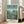 May include: Framed Studio Ghibli Ponyo movie poster displayed in bright living room corner next to beige sofa. Poster features red-dressed girl character standing on green cliff overlooking seaside village with blue ocean and distant islands. Japanese and English text at top, production credits at bottom. Light wood frame on white wooden floor against white walls with large windows.