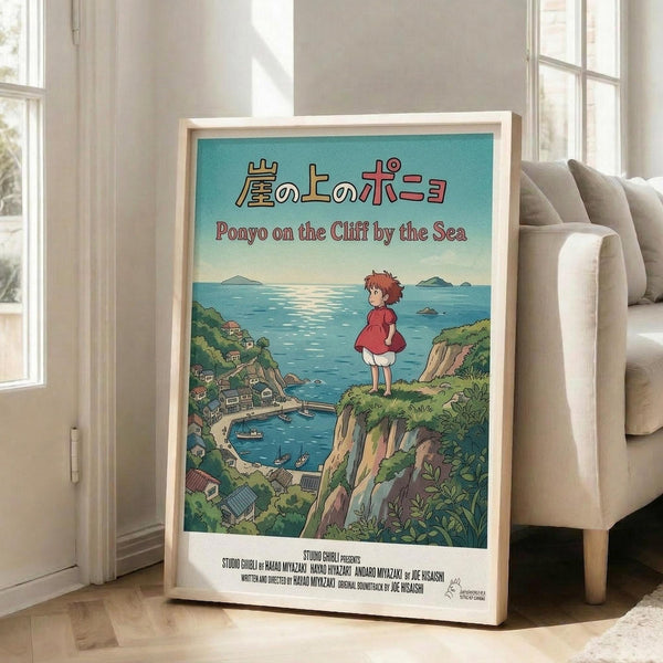 May include: Framed Studio Ghibli Ponyo movie poster displayed in bright living room corner next to beige sofa. Poster features red-dressed girl character standing on green cliff overlooking seaside village with blue ocean and distant islands. Japanese and English text at top, production credits at bottom. Light wood frame on white wooden floor against white walls with large windows.