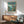 May include: Framed Studio Ghibli Ponyo movie poster hanging on white wall above wooden sideboard with lamp. Poster shows red-dressed girl on coastal cliff overlooking blue ocean with Japanese and English text. Mid-century modern furniture including teal upholstered chair with wood arms creates cozy living room setting with natural sunlight casting shadows.