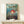 May include: Framed Studio Ghibli Ponyo movie poster displayed on light wood shelf against white wall. Features red-haired boy character swimming in ocean waves with coastal town backdrop. Japanese and English text visible. Small white wrapped gift with green sprig sits nearby. Warm natural lighting creates soft shadows.