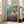 May include: Framed Studio Ghibli Ponyo movie poster leaning against white wall next to beige couch in bright living room with large windows. Poster shows red-haired Ponyo character swimming underwater in teal and green ocean scene with Japanese text and "Ponyo on the Cliff by the Sea" title. Natural wood frame against hardwood floors.