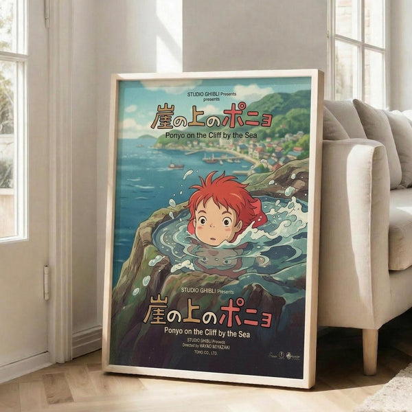 May include: Framed Studio Ghibli Ponyo movie poster leaning against white wall next to beige couch in bright living room with large windows. Poster shows red-haired Ponyo character swimming underwater in teal and green ocean scene with Japanese text and "Ponyo on the Cliff by the Sea" title. Natural wood frame against hardwood floors.
