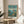 May include: Framed Studio Ghibli Ponyo movie poster displayed on light wood round side table next to beige upholstered chair. Poster shows red-haired child character standing on green cliff overlooking turquoise ocean with small coastal village below. Japanese and English text at top, credits at bottom. Natural lighting creates soft shadows on white wall behind.