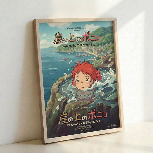 A framed Studio Ghibli movie poster for Ponyo on the Cliff by the Sea, featuring the red-haired character Ponyo emerging from splashing ocean water. The poster shows a coastal village scene with blue waters, green cliffs, and traditional buildings in the background. Japanese and English text appears at the top and bottom. The artwork is displayed in a light wood frame, leaning against a white wall on what appears to be a white surface.