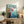 May include: Framed Studio Ghibli Ponyo poster displayed on rustic wooden sideboard in cozy living room setting. Poster shows red-haired boy jumping off coastal cliff into blue ocean water with Japanese text above. Natural wood frame, surrounded by fern plant, ceramic vase, stacked books, and cream upholstered chair with brown leather pillow.