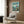 May include: Framed Studio Ghibli Ponyo movie poster mounted on wood-paneled wall above cream upholstered armchair with geometric patterned pillow. Poster shows red-haired child character overlooking coastal cliff town with turquoise ocean. Modern minimalist reading nook with brass floor lamp and dark round side table with open book.