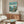 May include: Framed Ponyo movie poster hanging above wooden sideboard in modern living room. Poster shows red-dressed girl character overlooking seaside village from green cliff, with Japanese and English text on turquoise background. Room features cream curved chair, glass coffee table, and brass floor lamp against neutral walls.